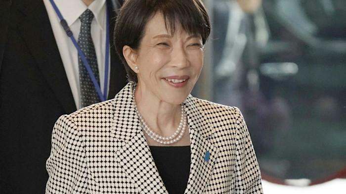 Sanae Takaichi smiles as she enters the prime minister's office wearing a patterned jacket and pearl necklace.