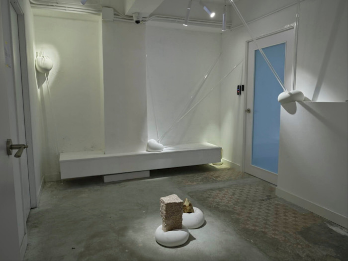 A contemporary art installation in a minimalist white room features sculptural objects, including a block and natural stone on white cushions, with translucent rods anchored to the walls.