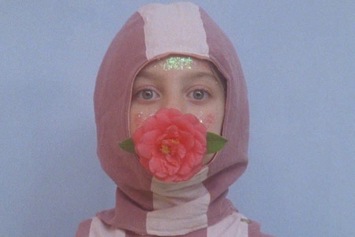 Still from an art film. A person in a pink and white hooded garment faces forward with a large pink flower covering their mouth. Their cheeks and forehead are decorated with glitter.