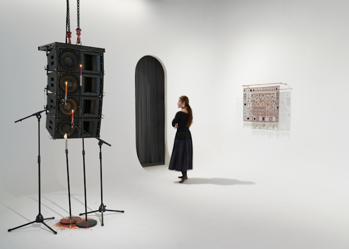 A woman walks around an art installation in a white-walled gallery. A large stack of loudspeakers are suspended from the ceiling by chains, over some mic stands which hold lit candles rather than microphones, all of them dripping red wax on the floor. 