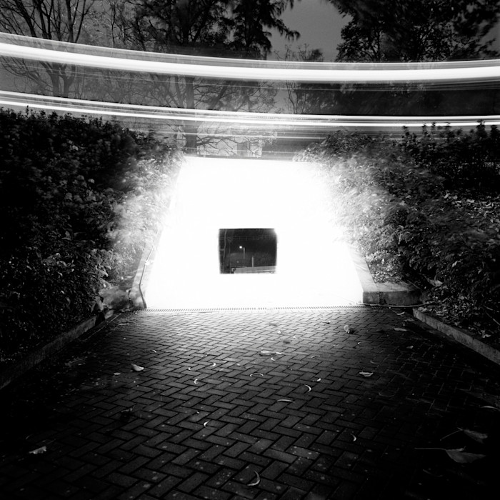 A black-and-white photograph of a tunnel entrance glowing with intense white light, with blurred light trails above and trees in the background.