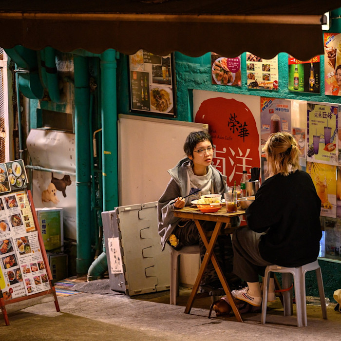 Two young women sit at a small table eating dinner and talking outside a restaurant decorated with colorful menus and posters.