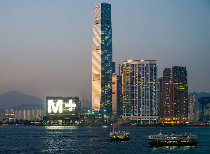 ICC International Commerce Centre and M+ museum are lit up at dusk beside Victoria Harbour, with ferries crossing the water in Hong Kong.