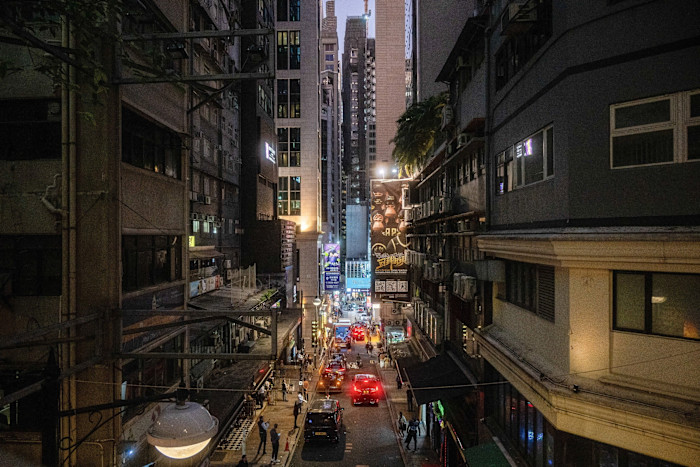 A narrow street in Lan Kwai Fong at dusk, lined with tall buildings, neon signs, cars, and groups of people.