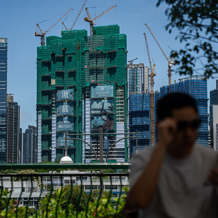 Deep Water Pavilia residential towers under construction, covered in green netting and surrounded by cranes, with a blurred person in the foreground.