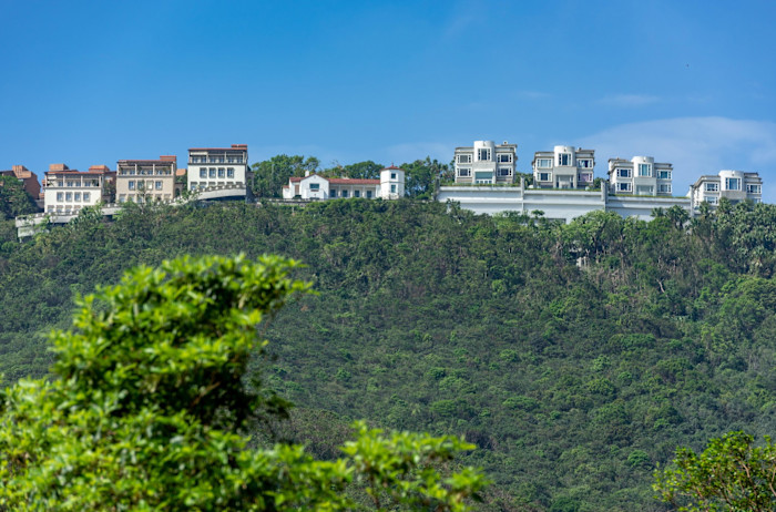 Luxurious homes line the top of a lush, green hillside under a clear blue sky along Peak Road in Hong Kong.