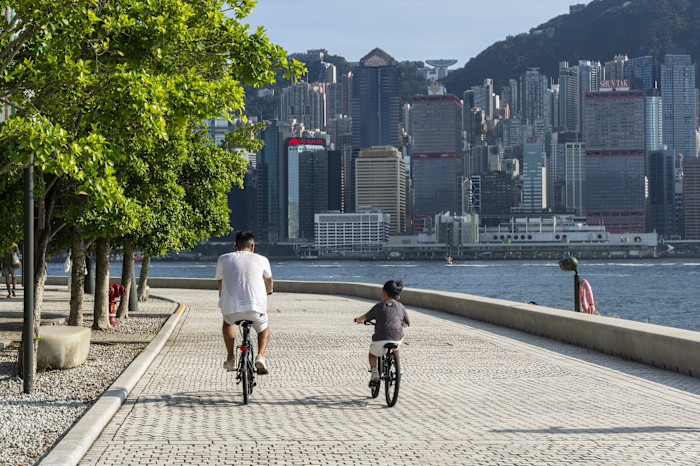 Two people ride bicycles along a waterfront path lined with trees, with Hong Kong skyscrapers visible across the water.