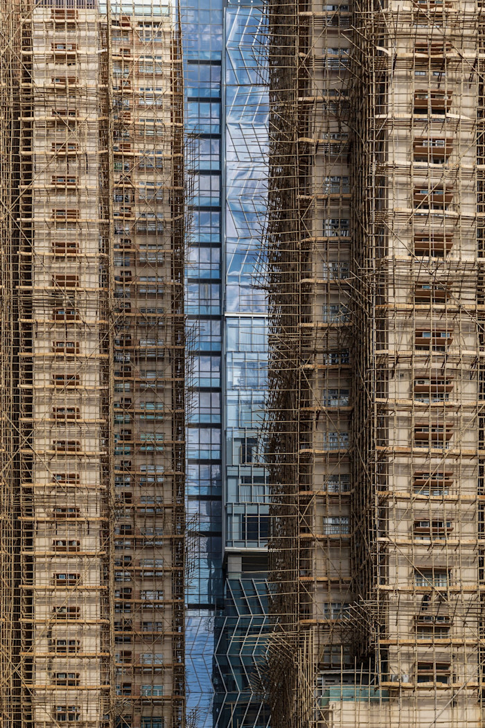 Residential buildings covered in dense bamboo scaffolding stand next to a modern glass building.