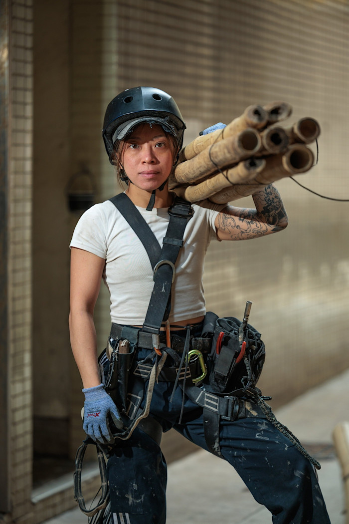 Daisy Pak stands near a construction site, wearing a helmet and safety gear, carrying several bamboo poles on her shoulder.