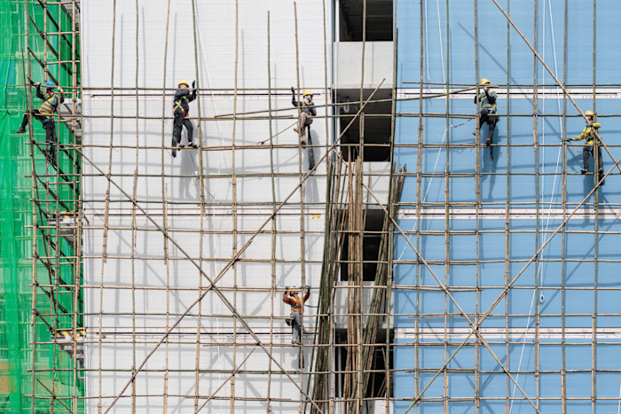 Workers wearing safety gear assemble bamboo scaffolding on the exterior of a high-rise building.