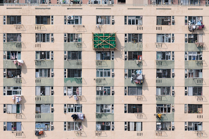 A bamboo scaffolding platform with green netting is attached to the exterior of a residential building among rows of windows and laundry.