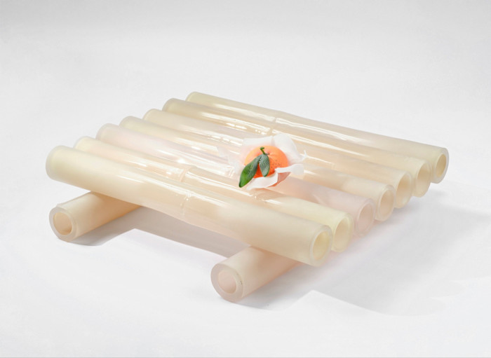 A sculptural arrangement of nine translucent glass tubes, shaped like bamboo, forms a raft, with a realistic glass mandarin and leaves resting on top.