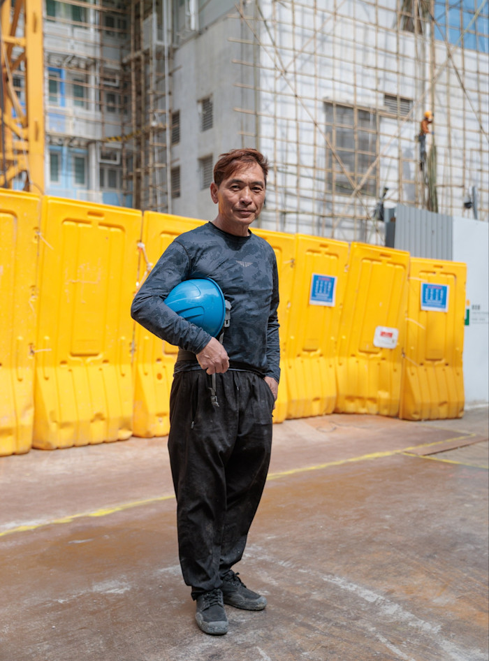 Kenny Lee stands outside a construction site holding a blue hard hat, with yellow barriers and scaffolding in the background.