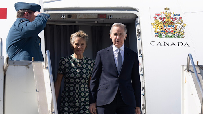 Prime Minister Mark Carney and Diana Fox Carney exit a Canadian government plane as a uniformed officer salutes.