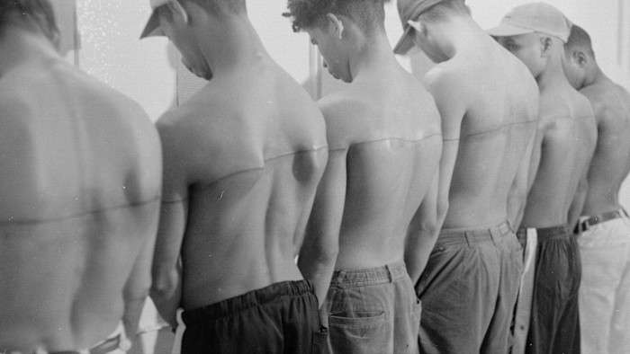 A black-and-white photograph, taken in Havana, Cuba in the 1990s, depicts six shirtless individuals standing in a row with a single tattooed line running horizontally across their backs.