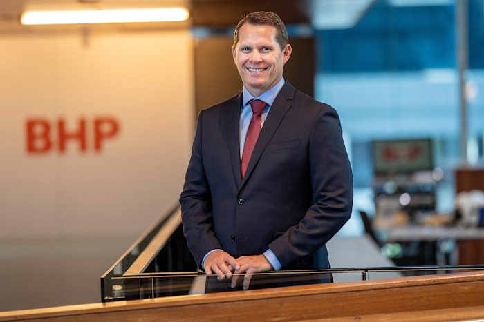 Brandon Craig stands in a business suit and red tie, smiling, in an office with a BHP sign in the background.