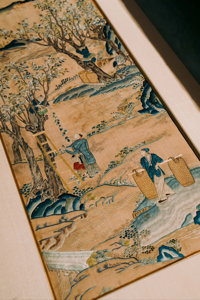 A detailed embroidered artwork showing figures gathering fruit from trees and carrying baskets in a landscape scene.