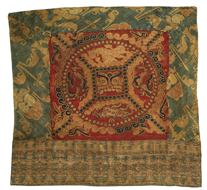 Photograph of a piece of embroidered silk fabric that serves as a pillow end. It depicts an animal mask in the centre of a circle, surrounded by smaller illustrations, in gold and dark blue on a red background. It dates from the Three Kingdoms period to Jin dynasty (220-420 CE). 