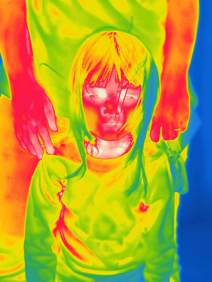 The outline of a child with her mother standing behind her. Both are bathed in swirls of bright red, orange and green. The image was created by using thermal imaging to evoke the invisible presence of radiation