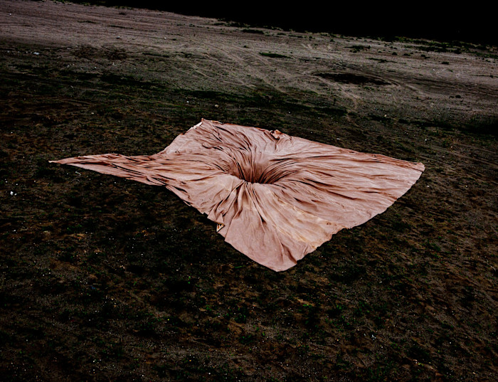 A piece of pink material is sucked into a hole in dark ground beneath it. The material sits in elegant folds around the hole, in sharp contrast to the rough earth around it 