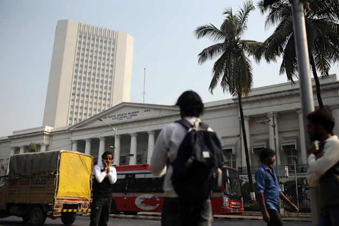 The Reserve Bank of India headquarters building in Mumbai stands behind palm trees, pedestrians, a red bus and a yellow delivery truck.