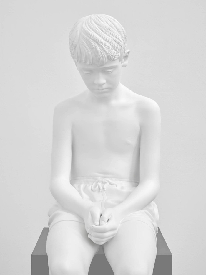 Photograph of a shiny white sculpture of a sad-looking, seated boy wearing just shorts, clasping his hands on his lap as he looks downwards. 