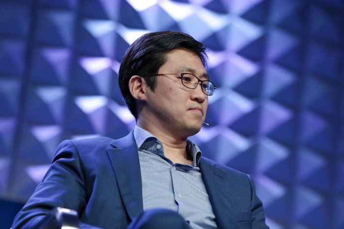Bom Kim listens on stage wearing a blue suit and glasses, with a geometric blue background behind him.