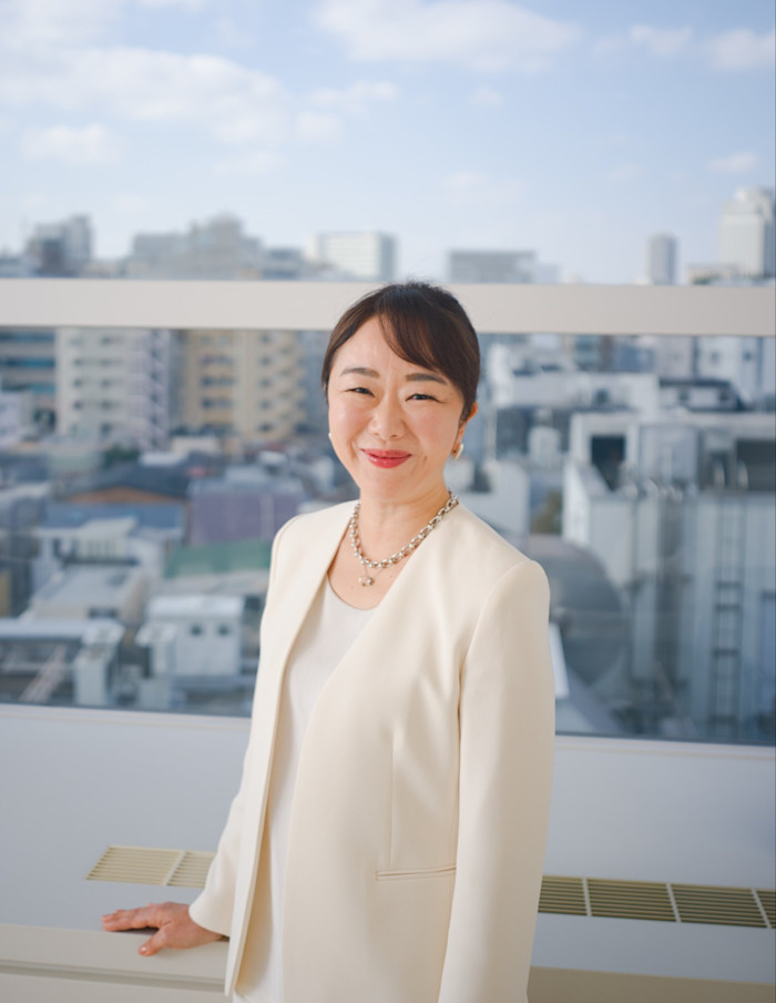 Maiko Todoroki smiles while standing in her office, with city buildings visible through the window behind her.