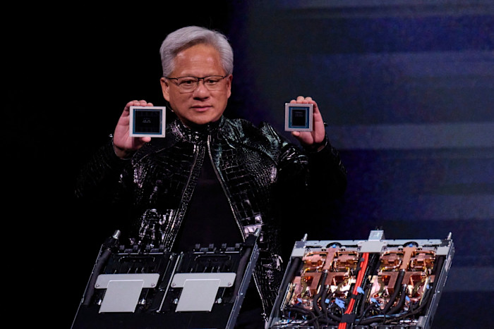 Jensen Huang holds up the Nvidia Rubin GPU and NVIDIA Vera CPU while presenting on stage, with computer hardware visible in front of him.