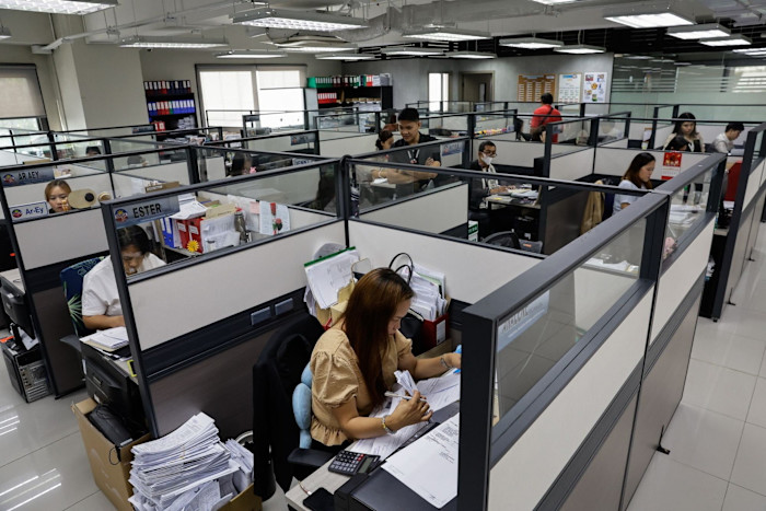 Employees of the Metropolitan Manila Development Authority work at desks in a busy office with cubicles and stacks of paperwork.