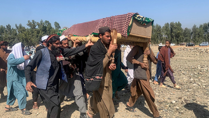 Mourners carry a coffin draped in patterned cloth during a funeral procession on rocky ground with others walking alongside.