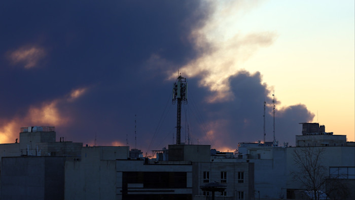 Smoke billows above city buildings in central Tehran after an air strike, with a communications tower in the foreground.