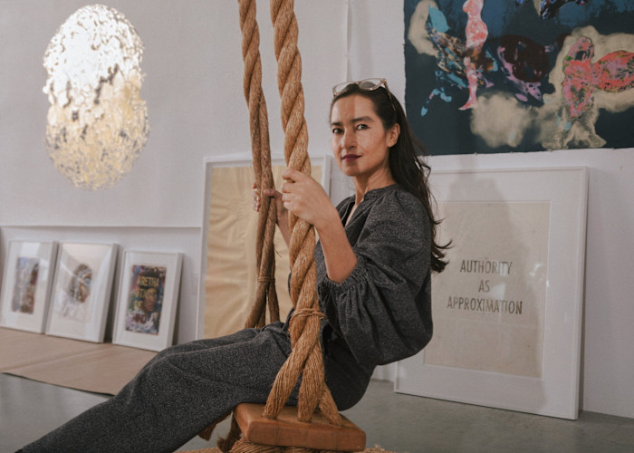 Shahzia Sikander sits on a wooden swing in an art studio, surrounded by framed artworks and a textured hanging light fixture.