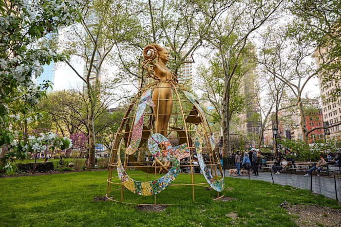 A large golden sculpture by Shahzia Sikander stands in a park, featuring a female figure with intricate hair inside a dome-like structure adorned with colourful mosaic patterns.