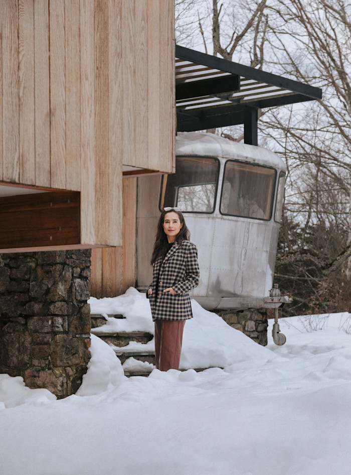 Shahzia Sikander stands in the snow outside a wood-panelled house, next to a 1950s-style silver caravan which appears to have been parked under a metal shelter but which is actually built into the structure of the house. 