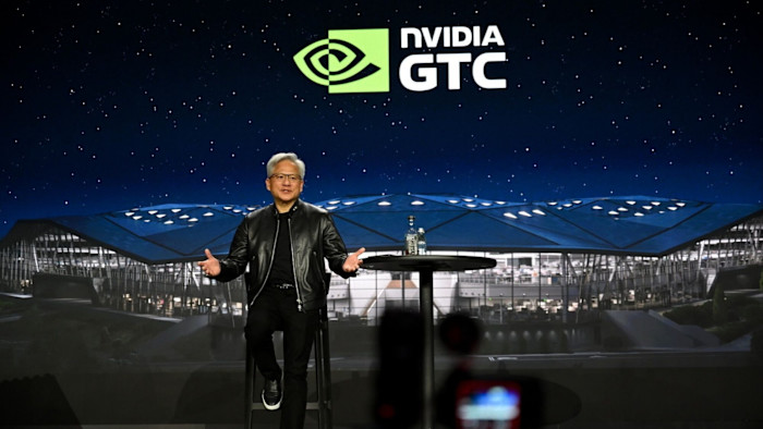 Jensen Huang speaks on stage at the Nvidia GTC conference, sitting at a table with a bottle of water. The Nvidia GTC logo is displayed behind him.