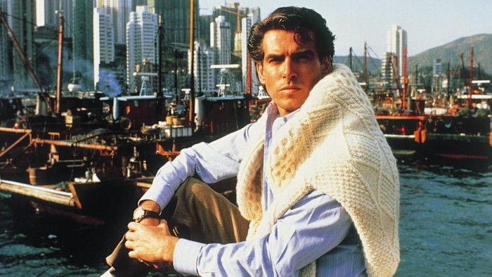Pierce Brosnan sits with a serious expression, draped in a cream-colored knitted sweater over a blue shirt against a backdrop of a bustling Hong Kong harbour, with boats and tall city buildings in the background, in a film still from ‘Noble House’